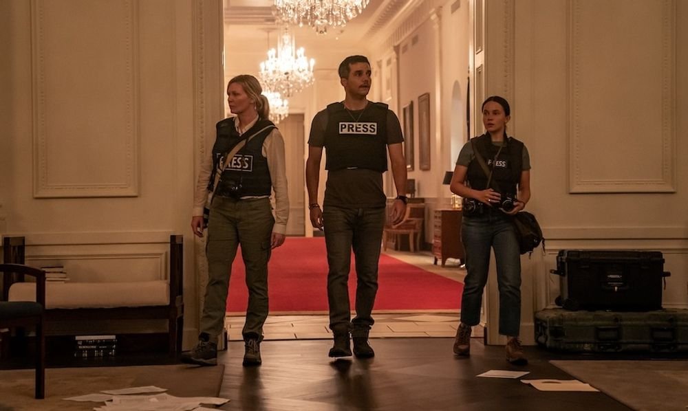 Três pessoas vestindo coletes de a prova de balas identificando-os como membros da impressa, dentro de um salão paredes brancas a frente de um corredor com tapete vermelho.