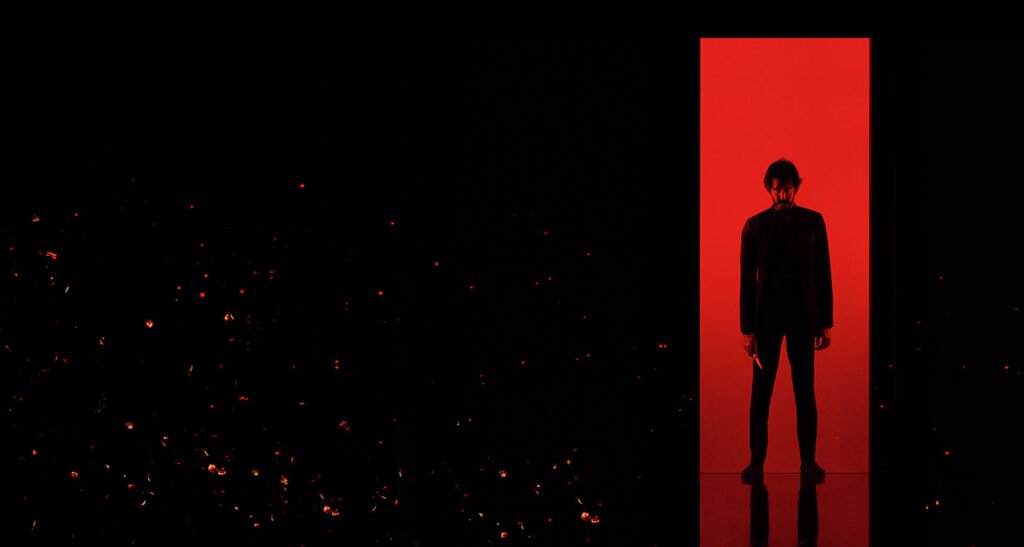 Fundo preto com faixa vermelha, representando um elevador, com a silhueta de um homem de preto segurando uma faca, no pôster de Monkey Man